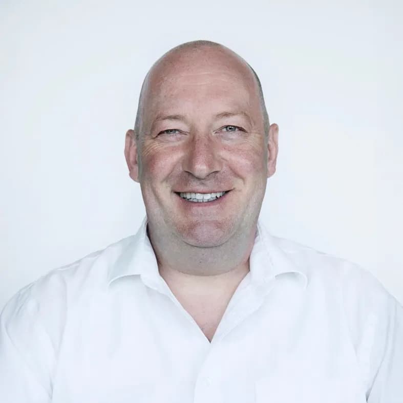 Er trägt ein weisses Hemd und lacht herzlich vor hellem Hintergrund.