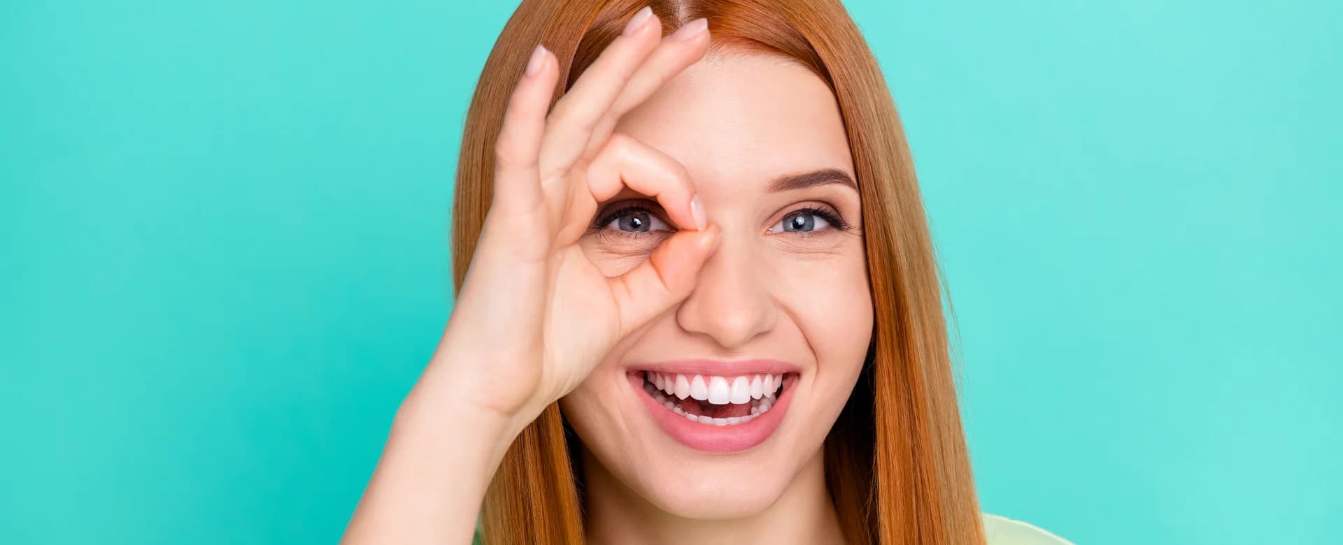 Junge Frau mit roten Haaren lächelt und formt mit Daumen und Zeigefinger ein ‘Okay’-Zeichen vor einem Auge, vor türkisfarbenem Hintergrund.