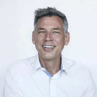 Er lächelt in die Kamera, trägt ein weisses Hemd und hat graues, seitlich frisiertes Haar.