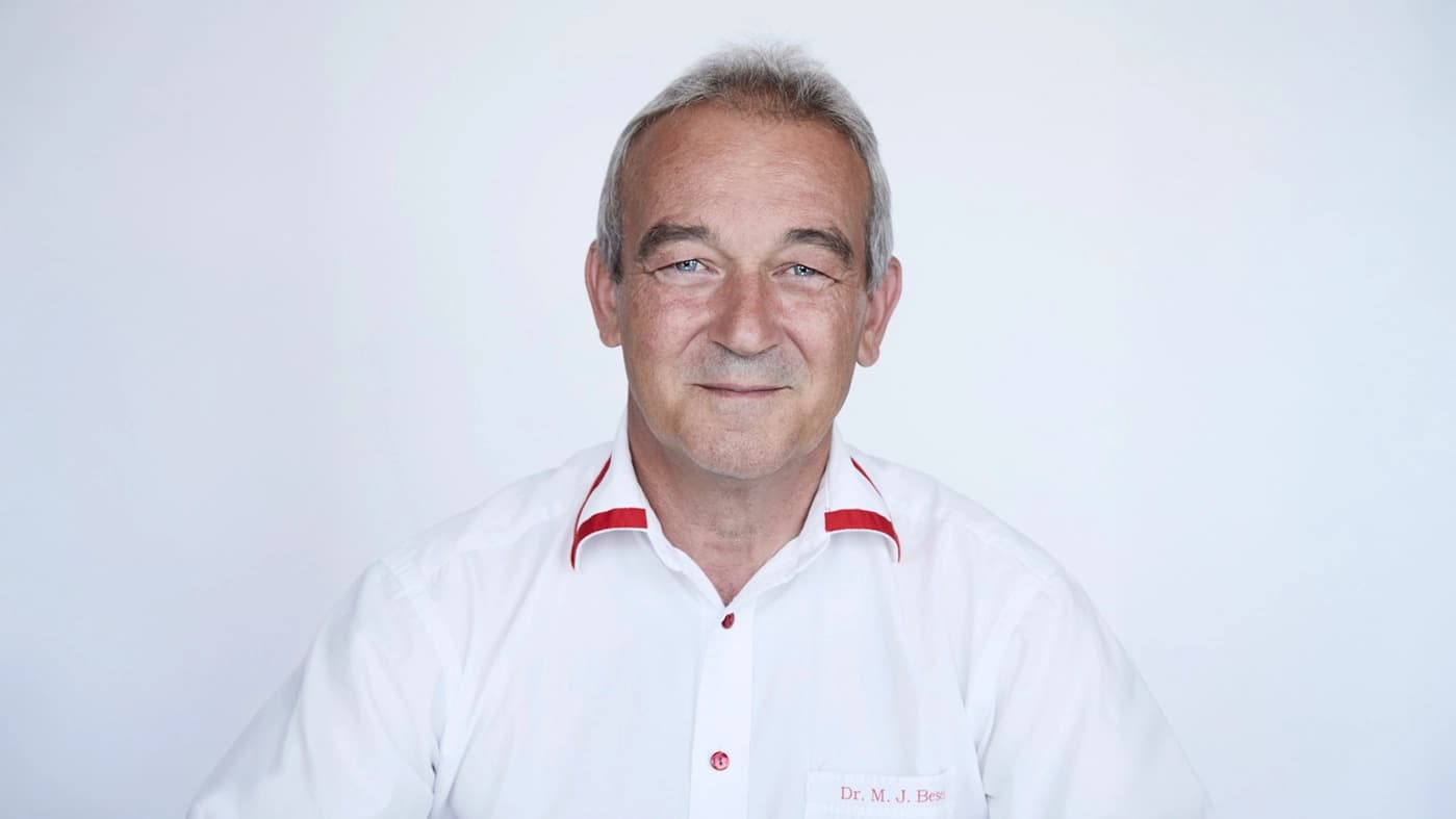 Er lächelt freundlich in die Kamera und trägt ein weisses Hemd mit roten Akzenten.