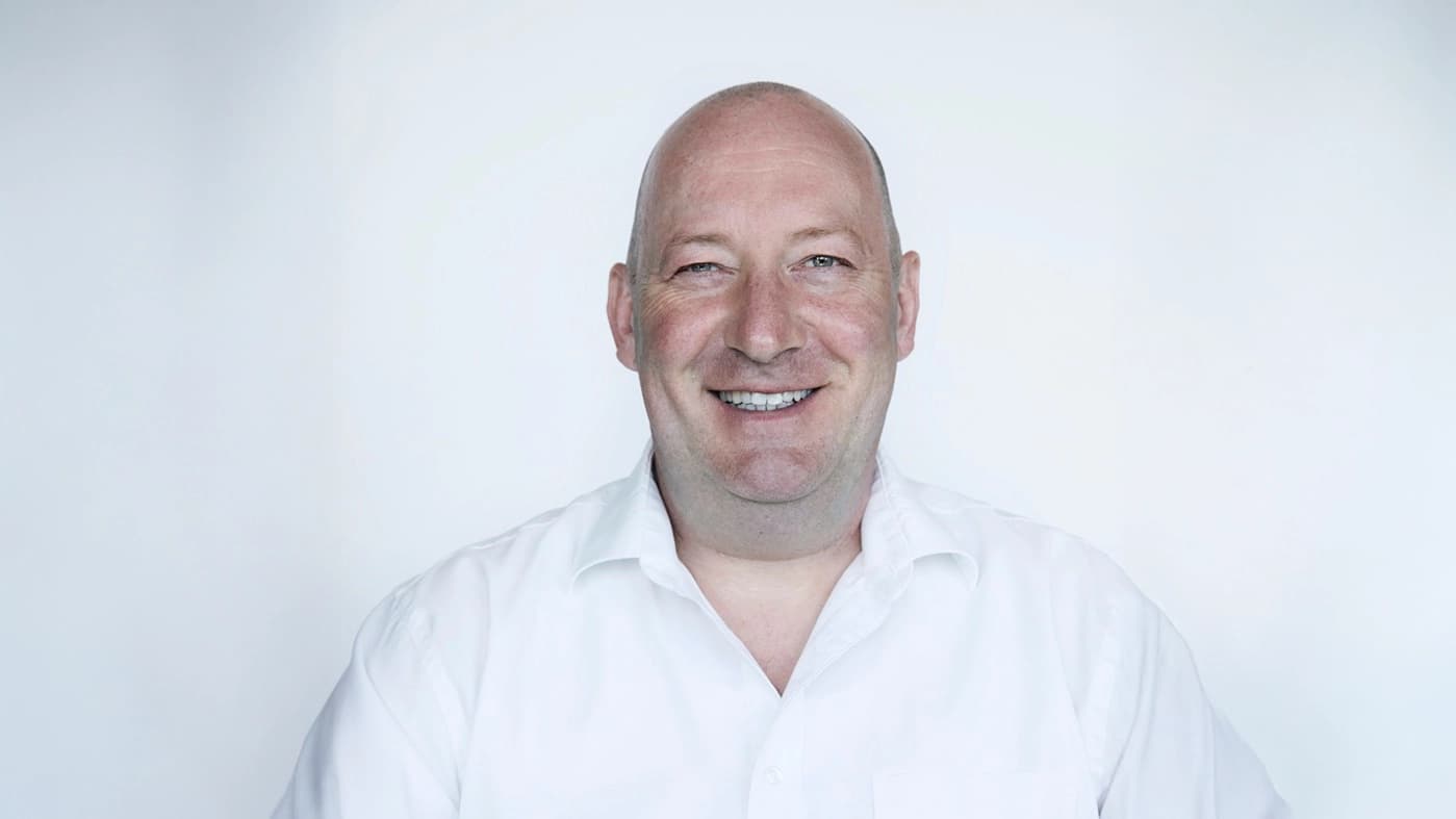 Er trägt ein weisses Hemd und lacht herzlich vor hellem Hintergrund.