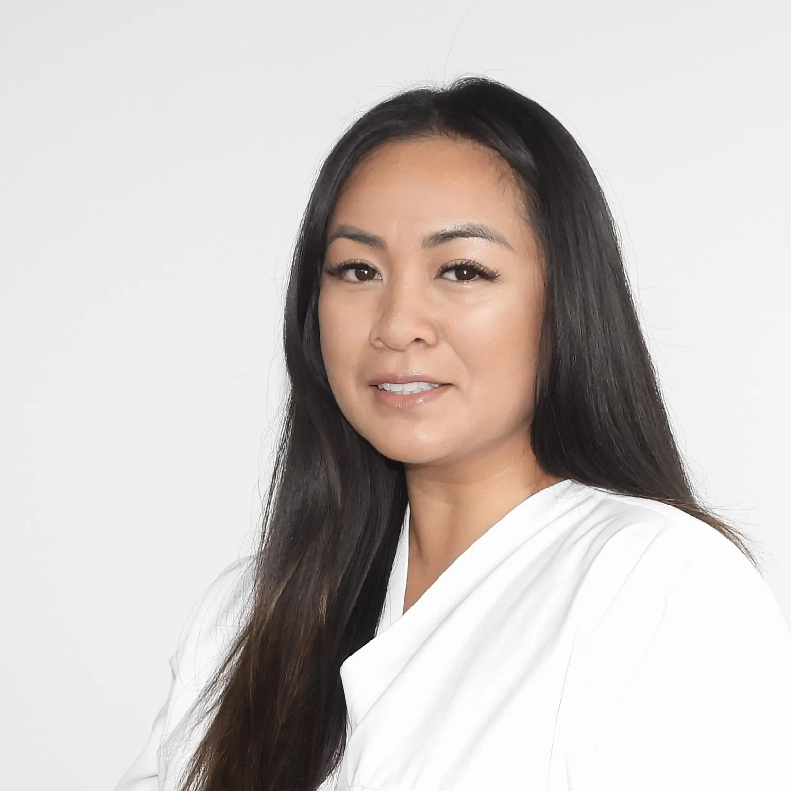 Sie hat lange, glatte, dunkle Haare, trägt Weiss und wirkt ruhig und professionell.