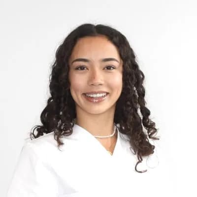 Ein Portraitbild. Sie trägt ein weißes Oberteil, eine Perlenkette und lächelt freundlich in die Kamera. Ihr dunkles, lockiges Haar fällt über die Schultern. Der Hintergrund ist hell und neutral.