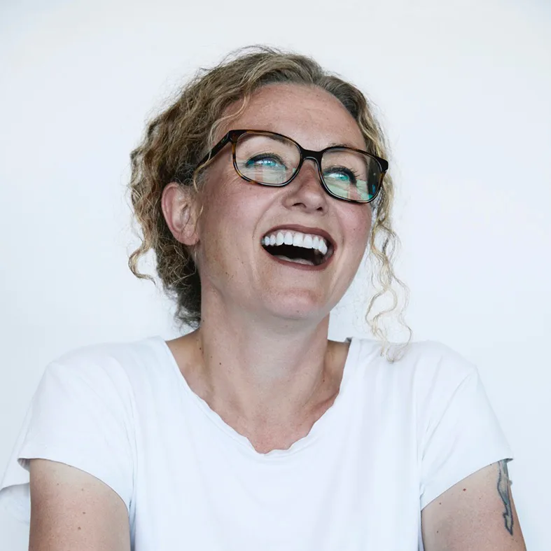 Sie lacht ausgelassen und trägt eine Brille sowie ein weisses Oberteil.