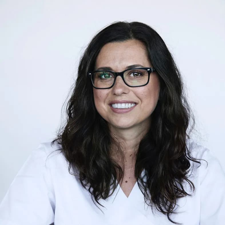 Sie trägt eine Brille, hat dunkle, wellige Haare und lacht herzlich in die Kamera.