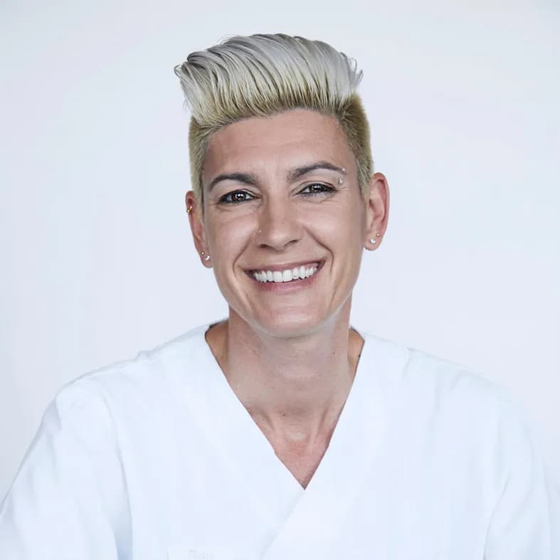 Sie lächelt breit, hat kurz geschnittenes Haar mit heller Strähne und trägt ein weisses Hemd.