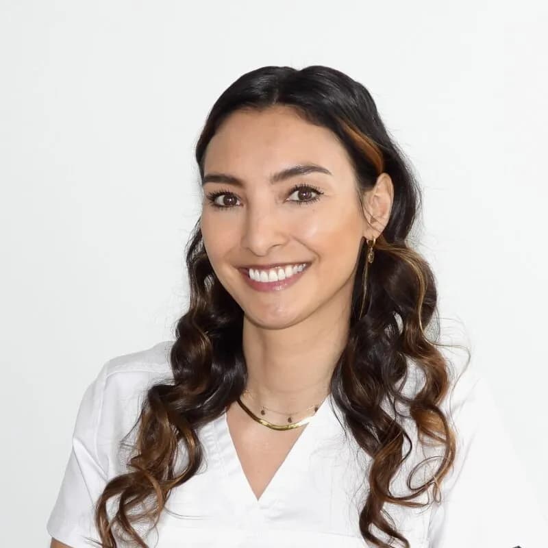 Sie lächelt herzlich, hat dunkle, gelockte Haare und trägt ein weisses Oberteil.